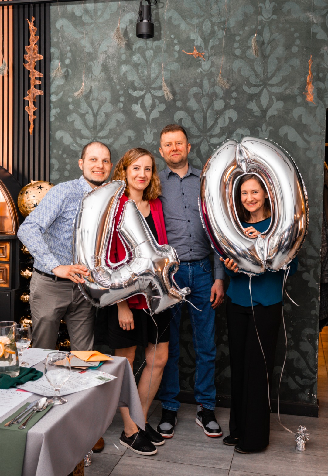 Grupa czterech osób świętuje urodziny, trzymając srebrne balony w kształcie cyfr '4' i '0', na tle dekoracyjnej ściany i stołu z zastawą.