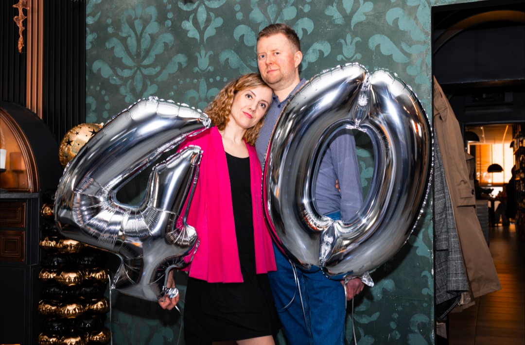 Para pozuje z srebrnymi balonami w kształcie cyfr '4' i '0' na tle wzorzystej ściany i fragmentu wnętrza lokalu.