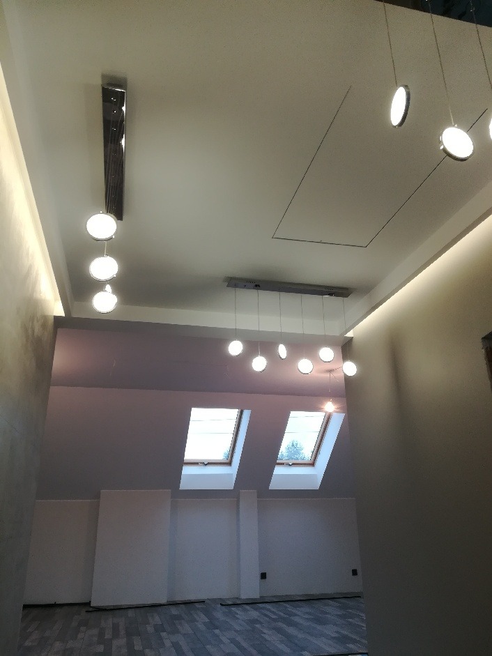 Wnętrze poddasza z dwoma oknami dachowymi, wykończone w minimalistycznym stylu, z szarą podłogą i białymi ścianami. Sufit zdobią nowoczesne lampy wiszące z okrągłymi kloszami.