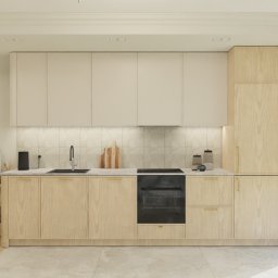 Magdalena Paryż - Jasna kuchnia z drewnianymi szafkami i beżowymi frontami, minimalistyczna lampa ścienna, płytki w tle, widok na salon z beżową sofą.