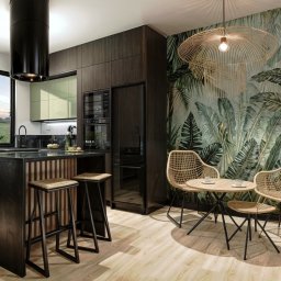 Sztukateria - projektowanie wnętrz w kamienicach w Szczecinie - Nowoczesna kuchnia z wyspą i wysokimi stołkami barowymi, połączona z jadalnią z okrągłym stołem i plecionymi krzesłami na tle tapety w liście tropikalne.