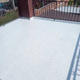 Jasnoszara posadzka z drobnymi plamkami na balkonie z metalową balustradą i ceglanym słupkiem. W tle zieleń i konstrukcja metalowa.