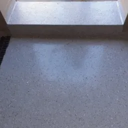 Szara, błyszcząca posadzka z drobnymi plamkami, z odpływem liniowym przy ścianie i progiem. Widok z góry, oświetlenie naturalne.