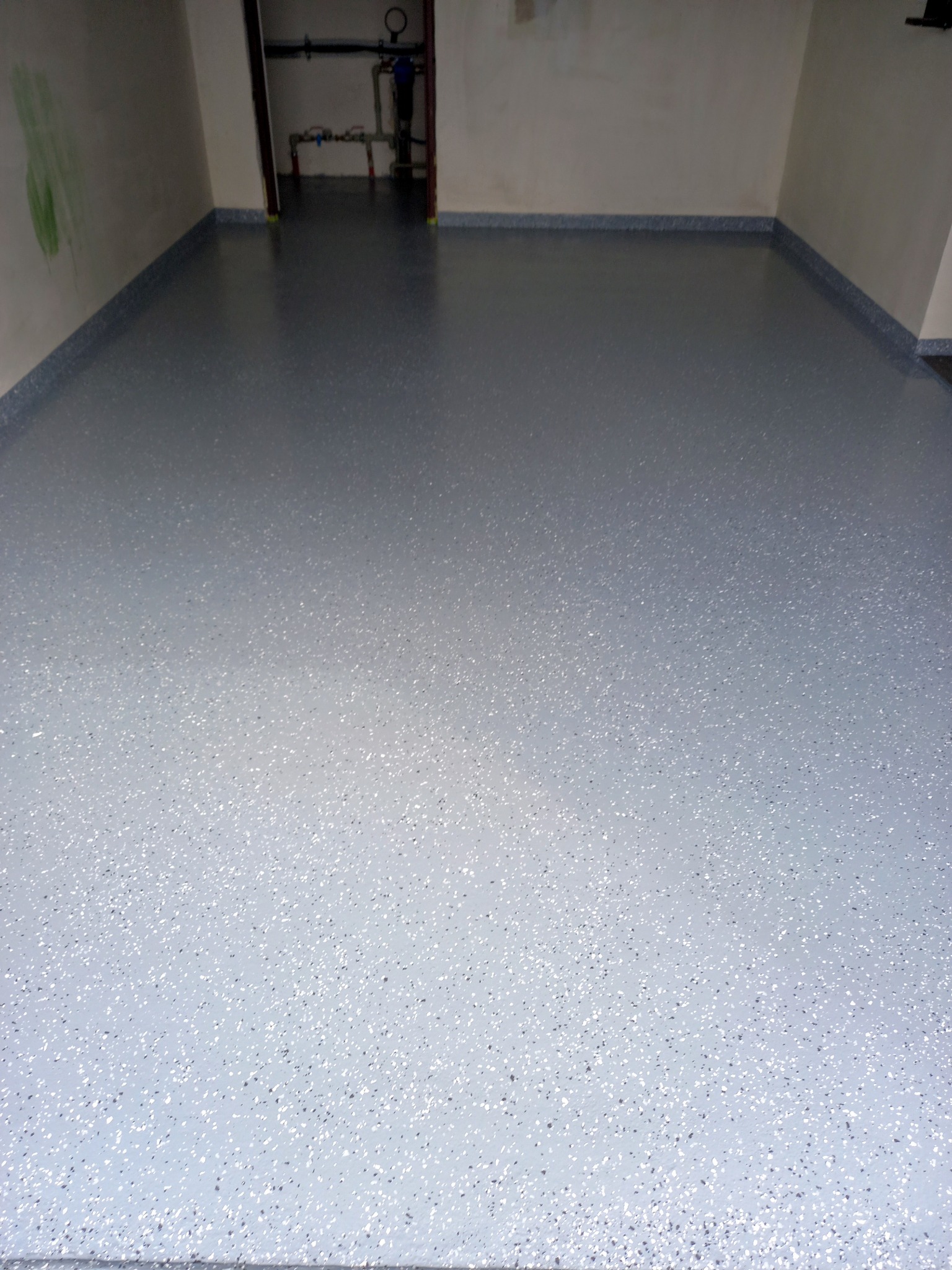 Szara, gładka powierzchnia posadzki żywicznej z delikatnym, jasnym wzorem w garażu. W tle widoczne instalacje i fragment ściany z zielonymi śladami.