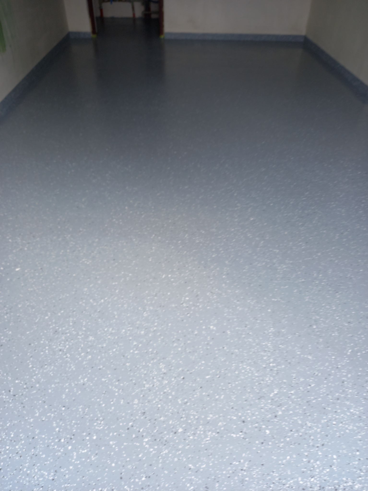 Jasnoszara, gładka posadzka z delikatnym, ciemnym wzorem, wykonana w pomieszczeniu z szarą listwą przypodłogową. Widoczny fragment otwartych drzwi w tle.