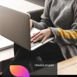 Osoba w szarym swetrze z żółtym akcentem pisze na laptopie. Widoczne ręce na klawiaturze. Ekran laptopa skierowany w stronę widza. Tło rozmyte.