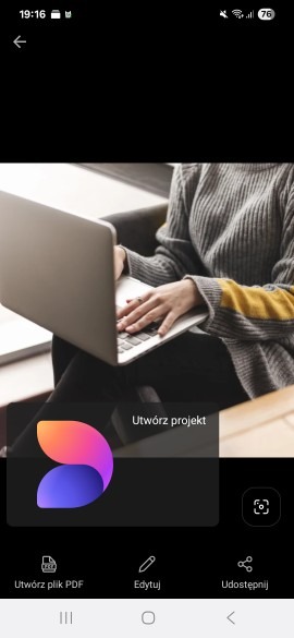 Osoba w szarym swetrze z żółtym akcentem pisze na laptopie. Widoczne ręce na klawiaturze. Ekran laptopa skierowany w stronę widza. Tło rozmyte.