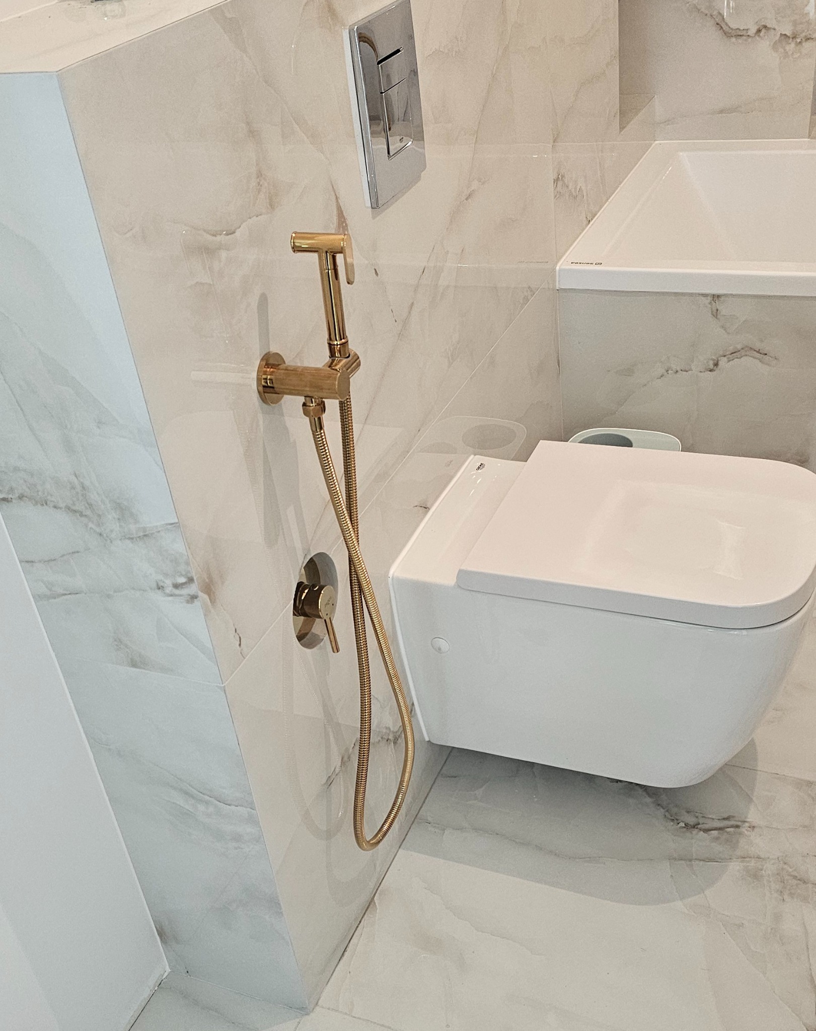 Nowoczesna łazienka z białą miską WC i złotym bidetem natryskowym na marmurowej ścianie. Minimalistyczny design, eleganckie wykończenie.