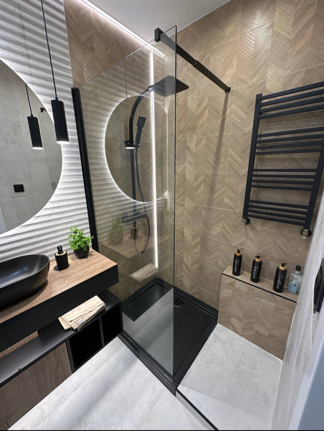 Nowoczesna łazienka z kabiną prysznicową typu walk-in, czarną armaturą i grzejnikiem drabinkowym. Umywalka nablatowa i oświetlenie LED dodają elegancji.