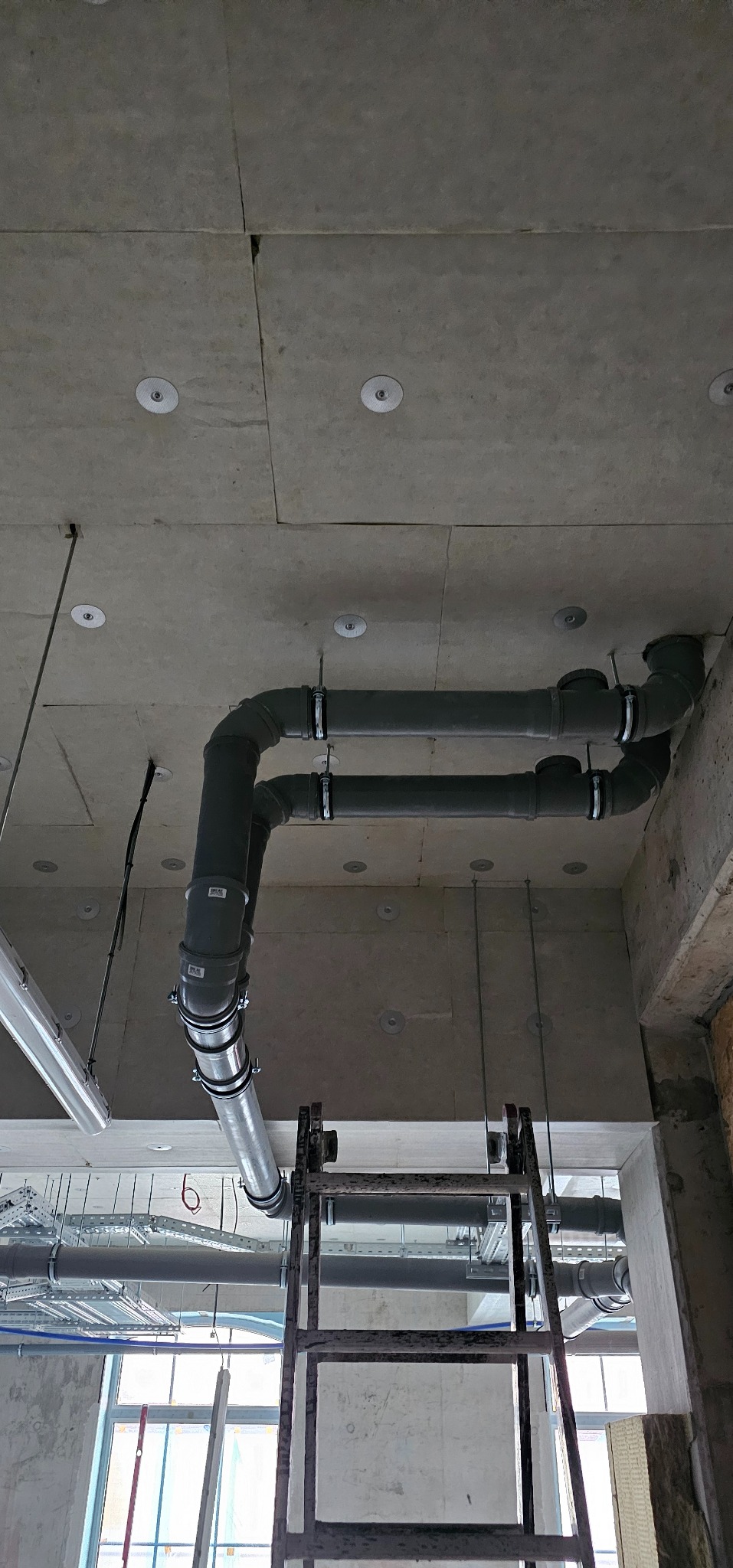 Szare rury wentylacyjne pod sufitem z widocznymi mocowaniami i fragmentem drabiny. Surowy stan wykończenia wnętrza, widoczne okablowanie i elementy konstrukcyjne.