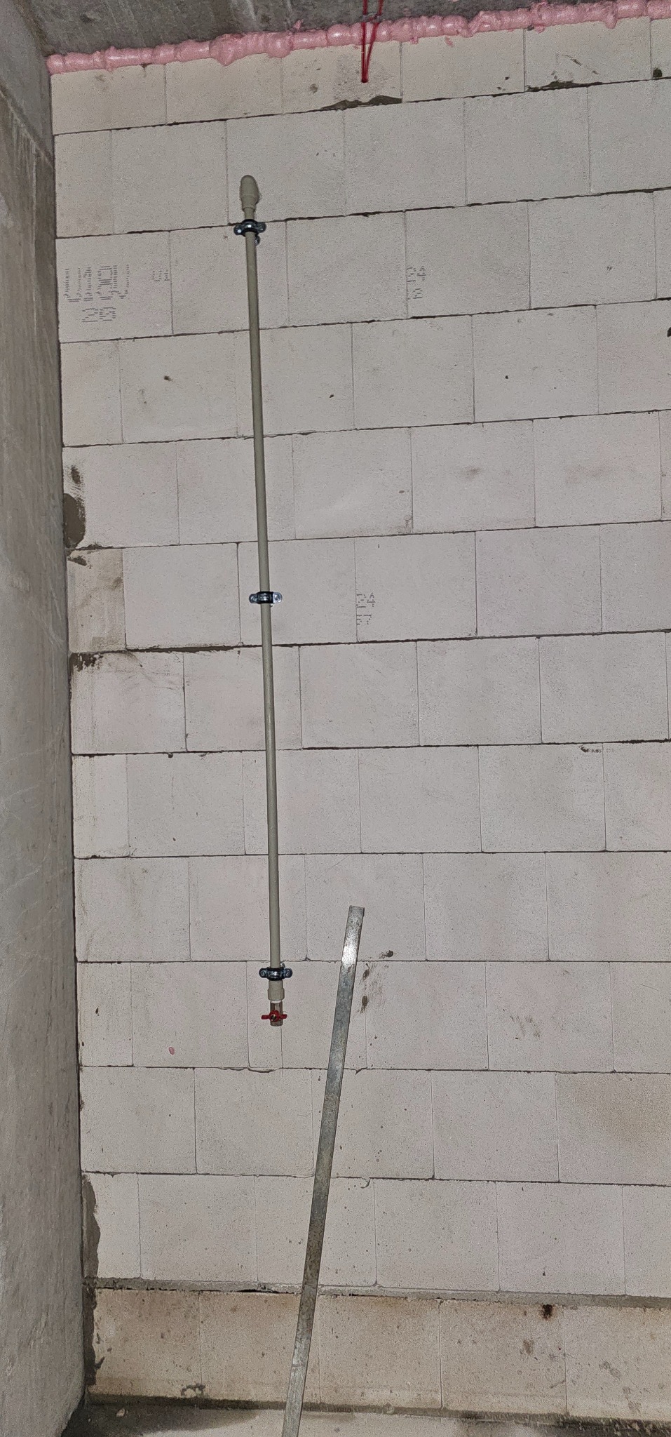 Szara rura pionowo zamocowana do ściany z białych bloczków, zabezpieczona klamrami. Widoczne różowe uszczelnienie na górze ściany i fragment metalowej listwy.