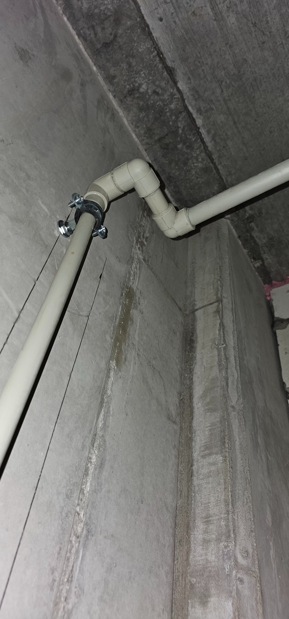 Szara rura z łącznikami przymocowana do surowej, betonowej ściany. Widoczne ślady wilgoci na ścianie. Ujęcie z dołu, podkreślające surowość wnętrza.