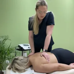 Kobieta w okularach i czarnym uniformie uciska dłonią plecy pacjentki leżącej na leżance z białym podkładem, w tle zielona ściana i roślina doniczkowa.