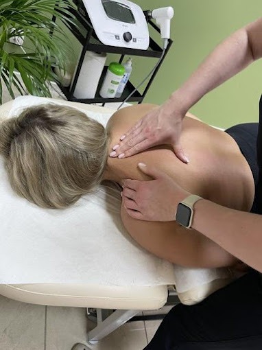 Masaż pleców pacjentki na leżance, widoczny sprzęt do fizykoterapii w tle, jasne wnętrze gabinetu.