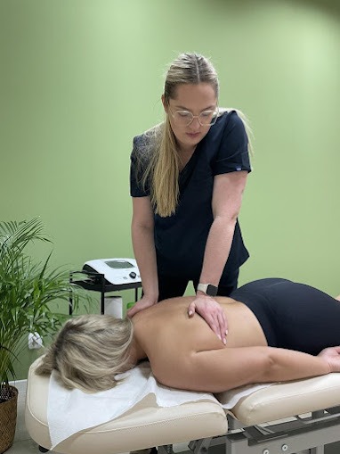 Kobieta w okularach i czarnym uniformie uciska dłonią plecy pacjentki leżącej na leżance z białym podkładem, w tle zielona ściana i roślina doniczkowa.