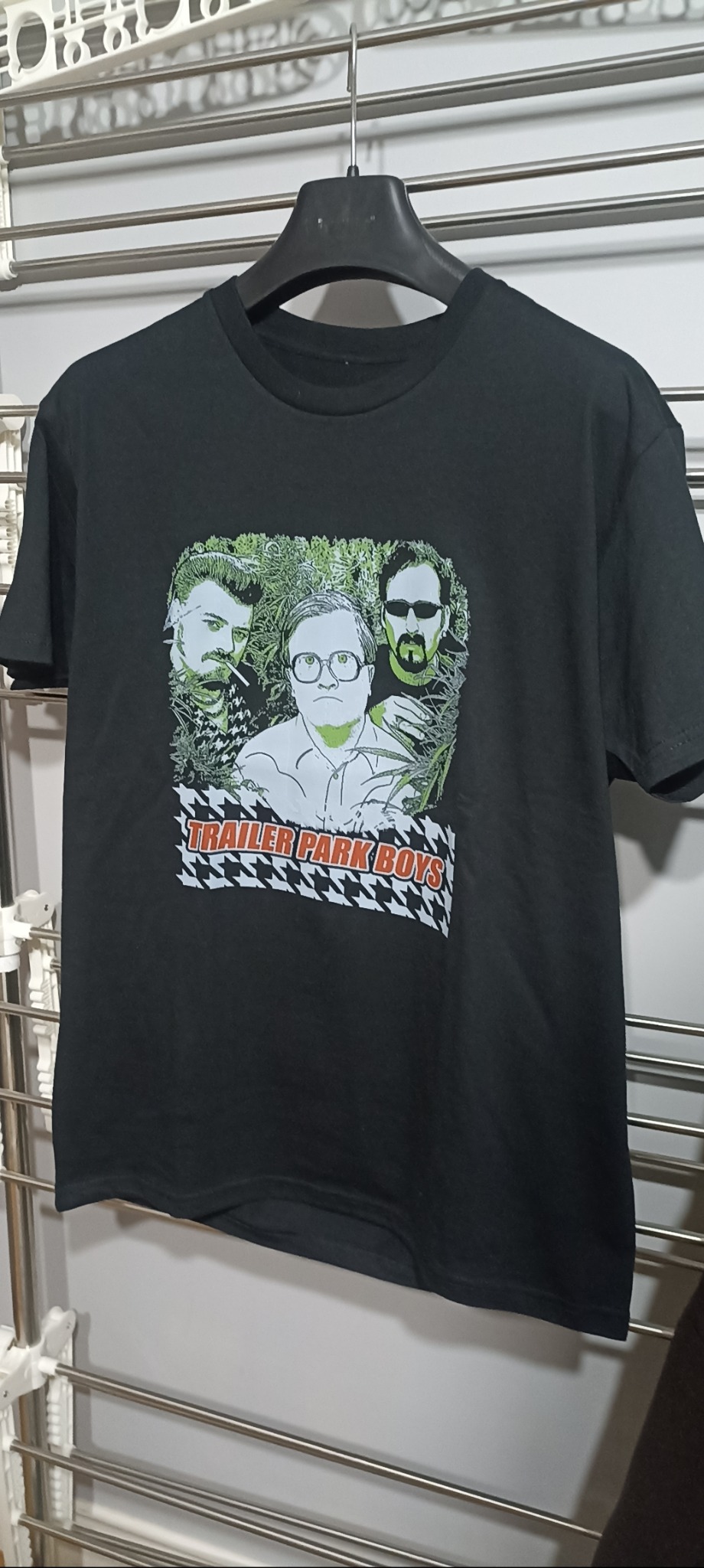 Czarna koszulka z nadrukiem postaci z serialu 'Trailer Park Boys' zawieszona na wieszaku, widoczny fragment suszarki do ubrań w tle.