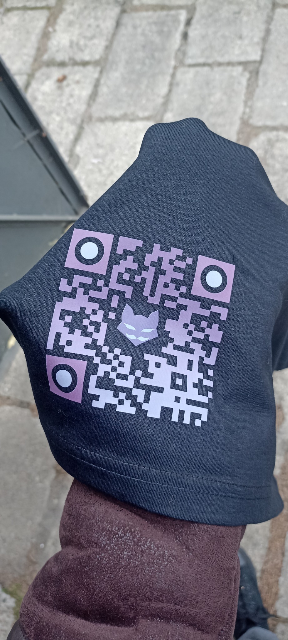 Ciemna koszulka z nadrukowanym fioletowo-białym kodem QR zawierającym w centrum stylizowaną głowę kota, trzymana w dłoni na tle chodnika.