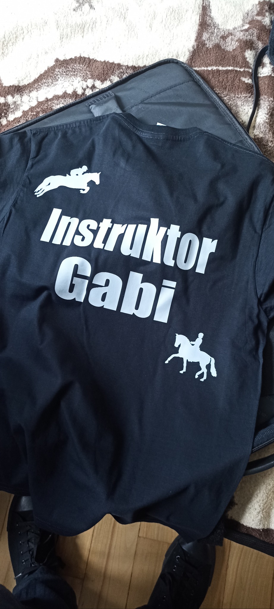 Czarna koszulka z białym napisem 'Instruktor Gabi' i grafiką jeźdźca na koniu, leżąca na podłodze obok czarnej torby i butów.
