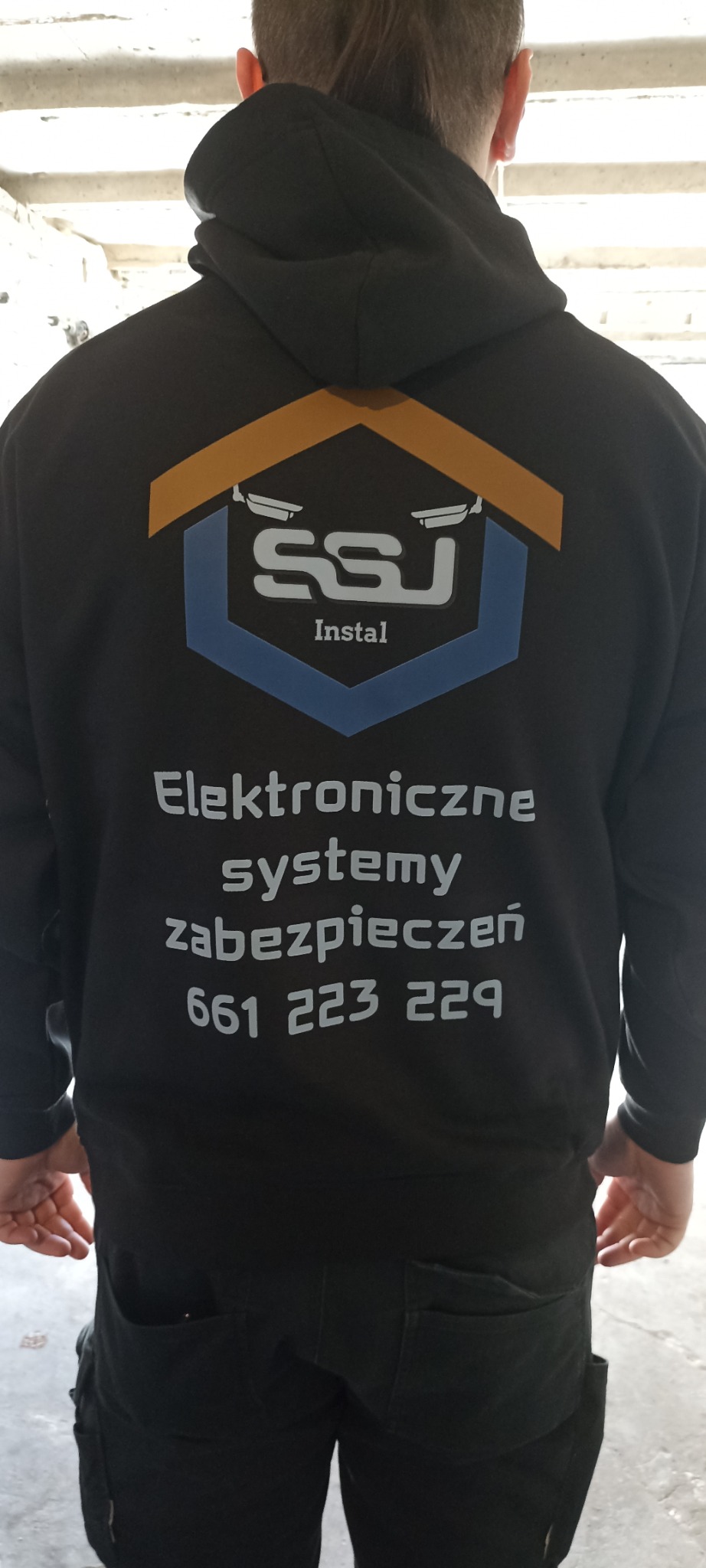 Czarna bluza z kapturem, na plecach logo SSJ Instal i numer telefonu do elektronicznych systemów zabezpieczeń, widok z tyłu na stojącą osobę.