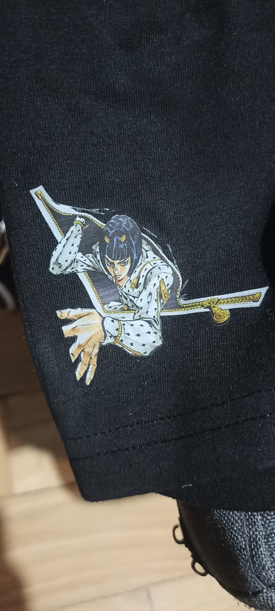 Czarna koszulka z nadrukiem postaci z anime, wychodzącej z rozpiętego zamka błyskawicznego. Widoczny fragment rękawa i szew.