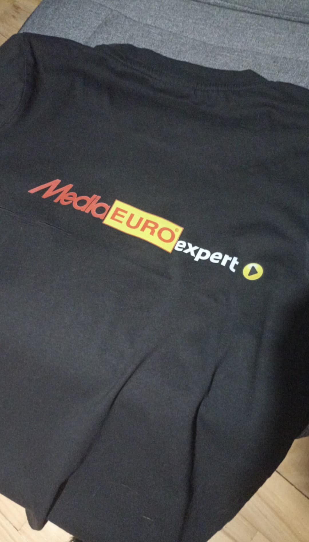 Czarna koszulka z nadrukowanym logo Media Expert, leżąca na szarym tle. Widoczne detale nadruku i faktura materiału koszulki.