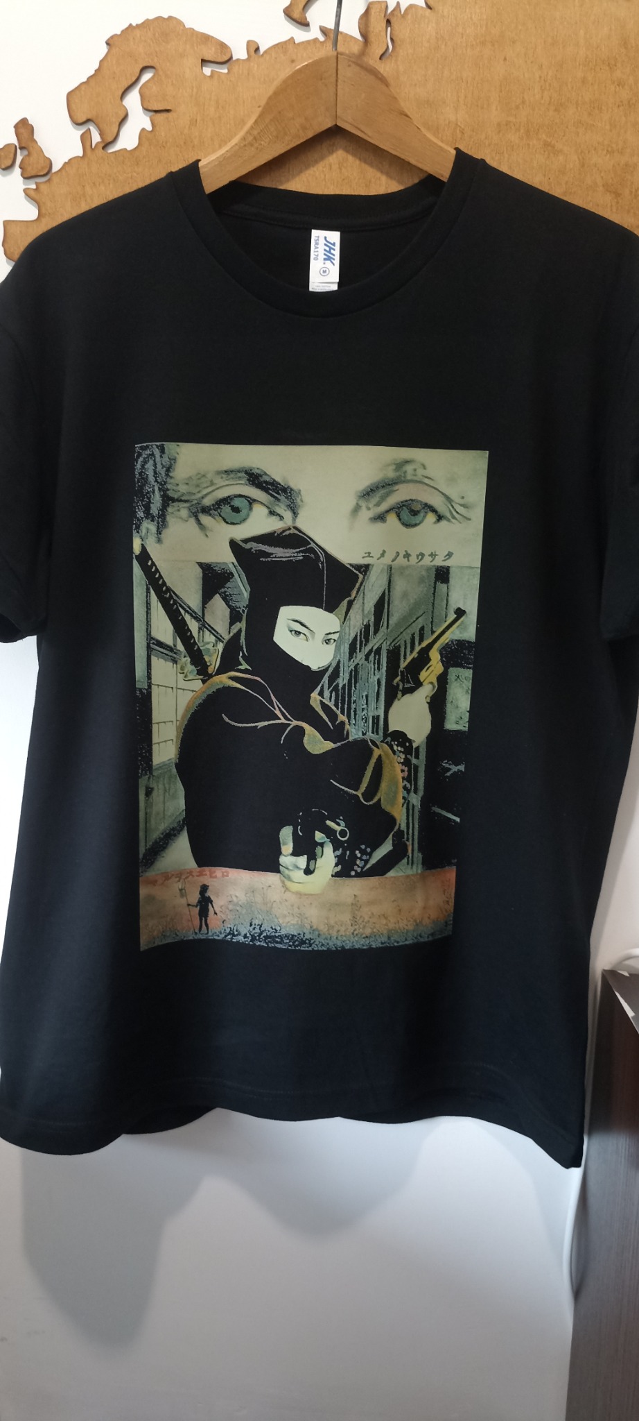 Czarna koszulka z nadrukiem w stylu anime, przedstawiającym postać ninja z bronią na tle miejskiego krajobrazu, zawieszona na drewnianym wieszaku.