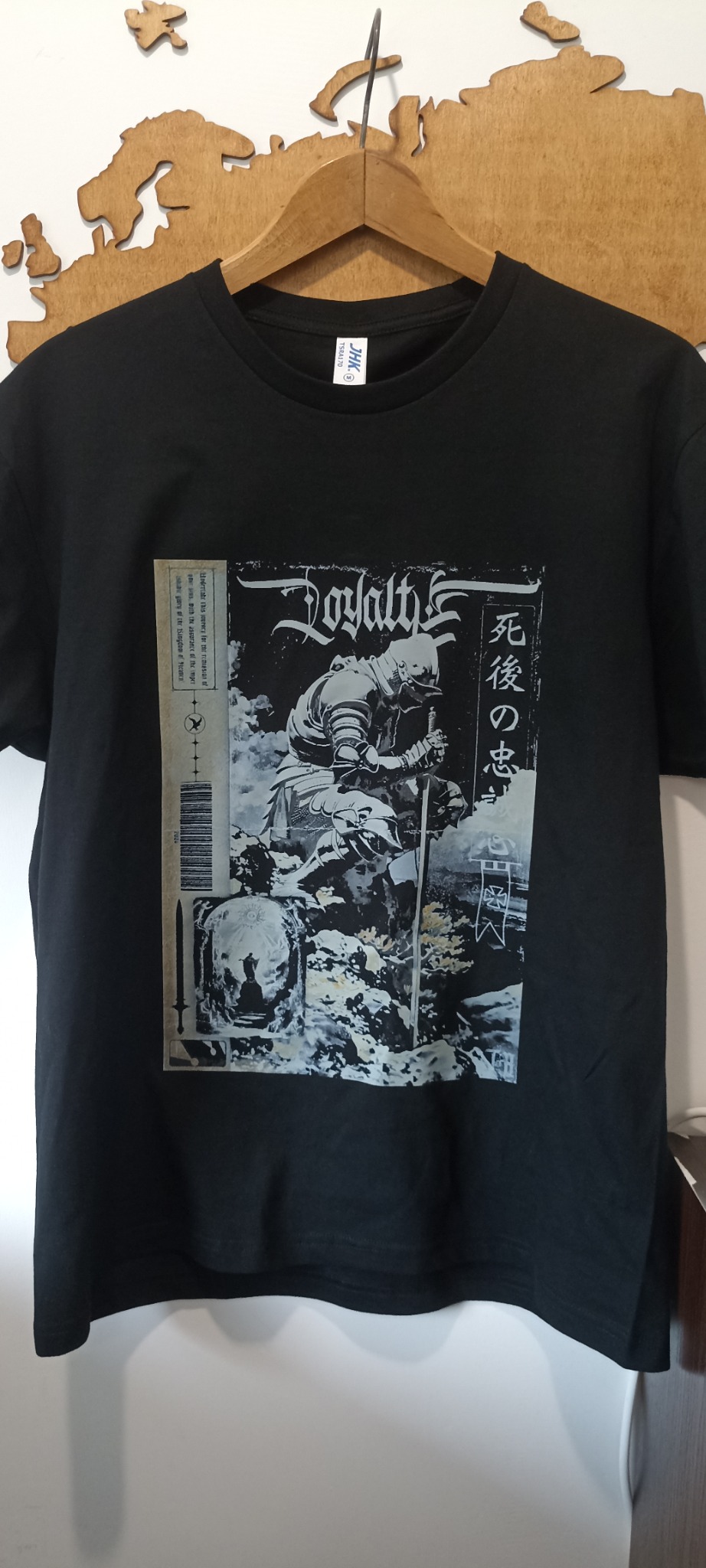 Czarna koszulka z nadrukiem w stylu gotyckim, przedstawiającym rycerza i japońskie znaki. Koszulka wisi na drewnianym wieszaku na tle mapy świata.