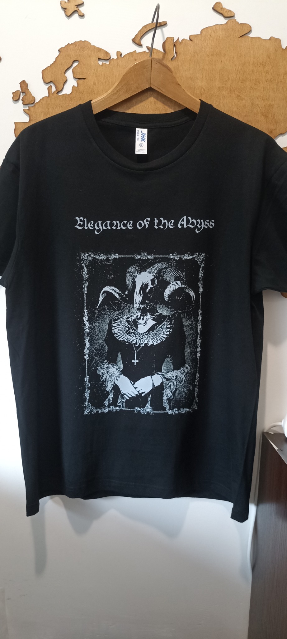 Czarna koszulka z nadrukiem w stylu gotyckim, wisząca na wieszaku. Grafika przedstawia postać w szatach z motywem czaszki i rogów. Napis: 'Elegance of the Abyss'.
