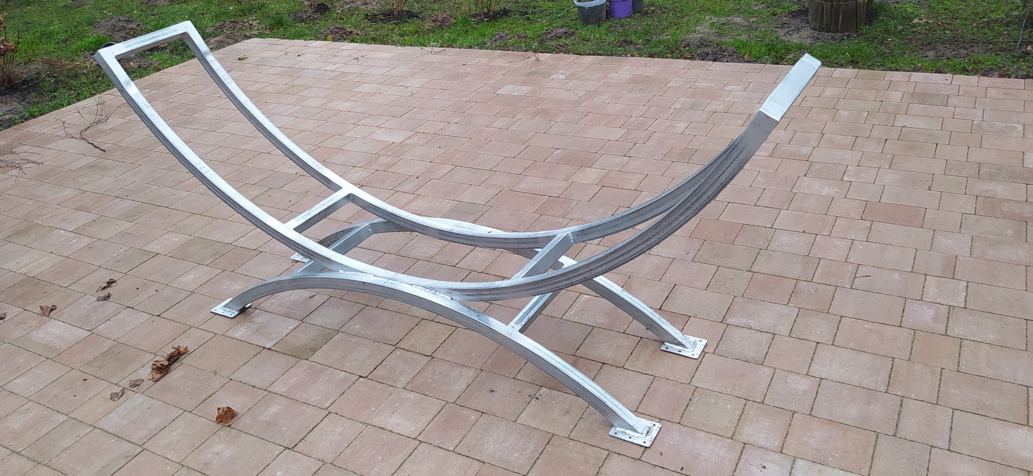 Metalowy, spawany stojak na drewno kominkowe, pomalowany na srebrno, ustawiony na brukowanej powierzchni. Widoczne ślady spawania i szlifowania.