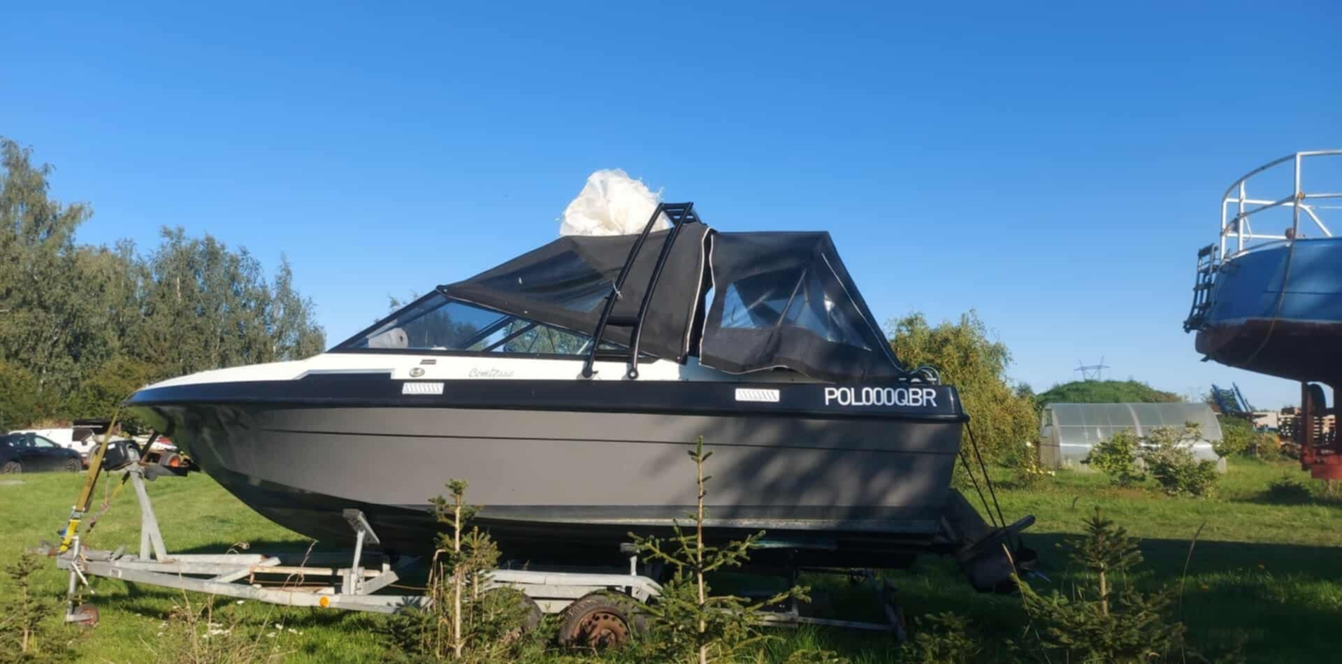 Szara łódź motorowa z czarnym zadaszeniem, zaparkowana na przyczepie na trawie, z numerem rejestracyjnym POL000QBR, w tle drzewa i szklarnia.