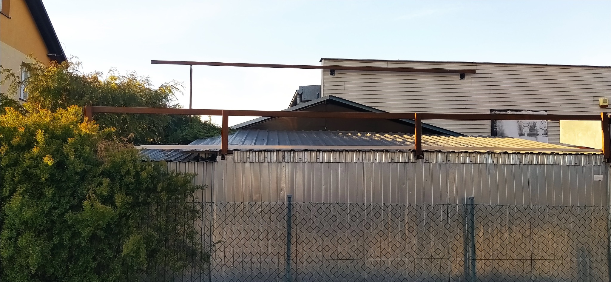 Metalowa konstrukcja w trakcie budowy, widoczna na tle blaszanej ściany i siatki ogrodzeniowej. Konstrukcja z belek, widoczne spawy. Częściowo zarośnięta krzewami.