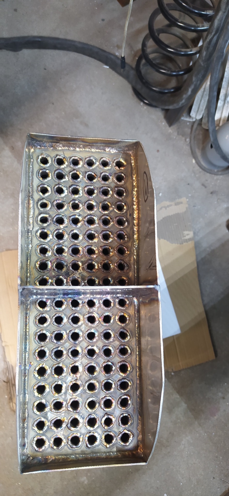 Metalowy element z otworami po obróbce, widoczne ślady spawania i szlifowania. Element wykonany z blachy, z regularnym układem otworów.