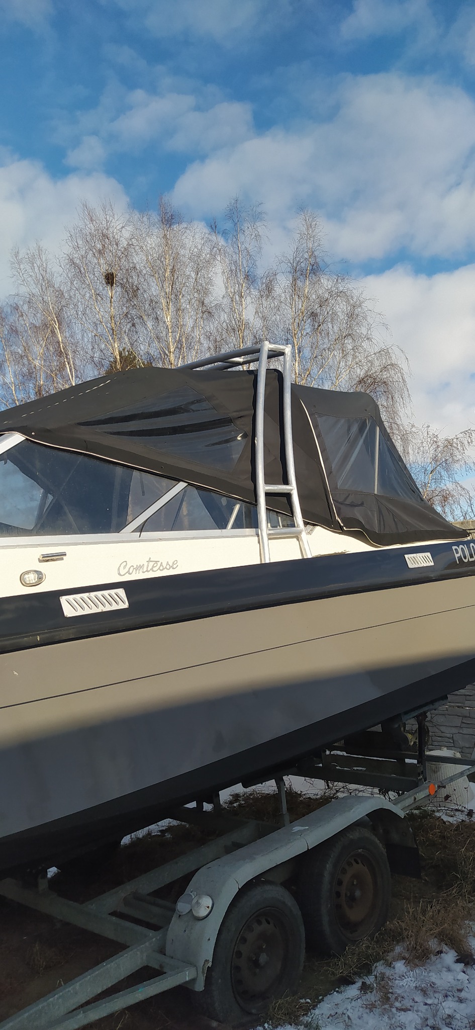 Szary jacht motorowy Comtesse na przyczepie, z czarnym pokrowcem i metalową konstrukcją, na tle zimowego krajobrazu z drzewami i błękitnym niebem.