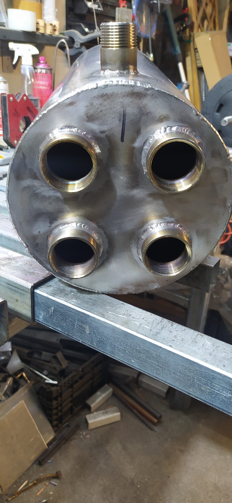 Spawany, metalowy zbiornik ciśnieniowy z czterema gwintowanymi otworami, umieszczony na metalowej konstrukcji w warsztacie. Widoczne ślady szlifowania.