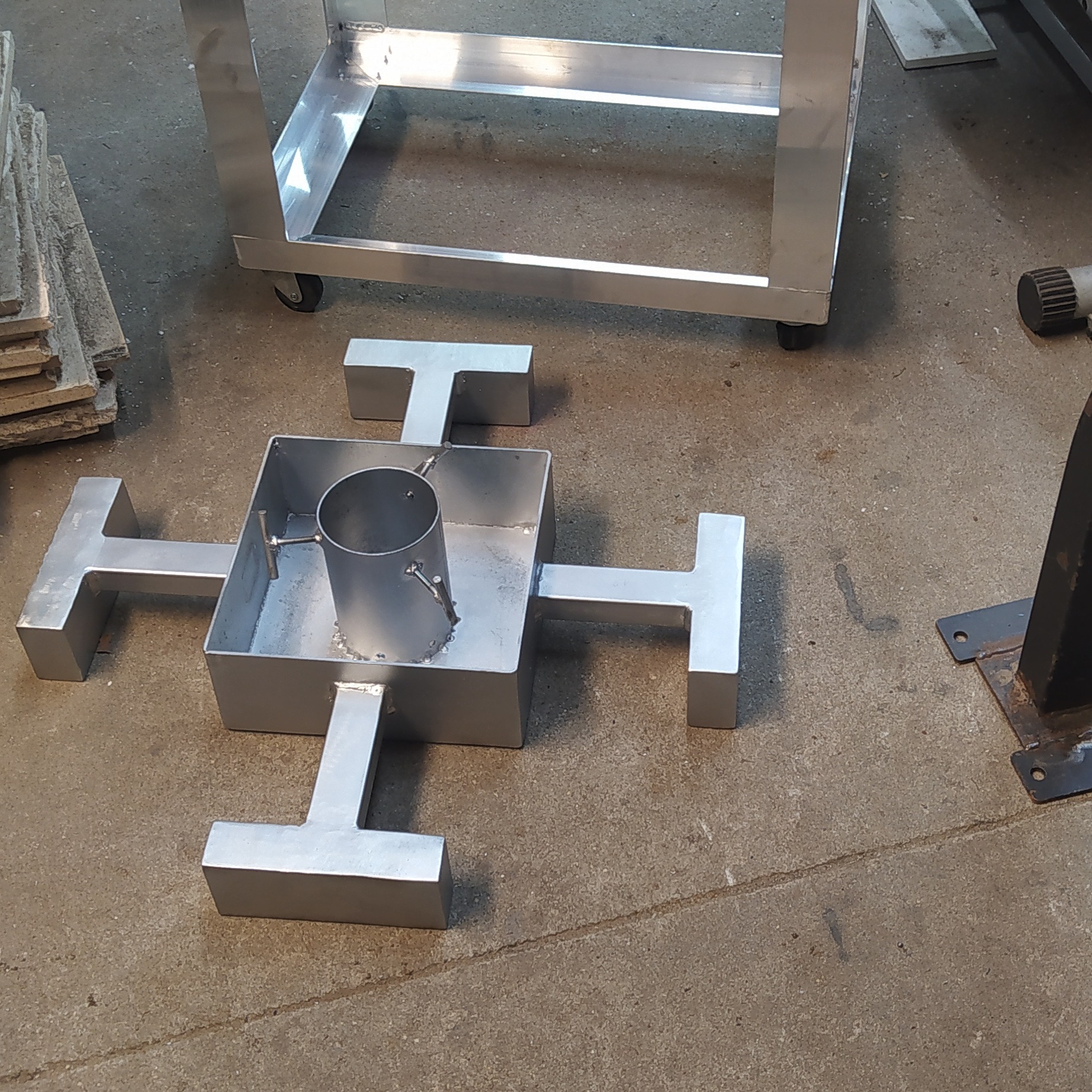 Metalowa konstrukcja spawana z rur i profili, w kształcie krzyża z centralnym cylindrem, stojąca na betonowej posadzce. Widoczny wózek warsztatowy w tle.