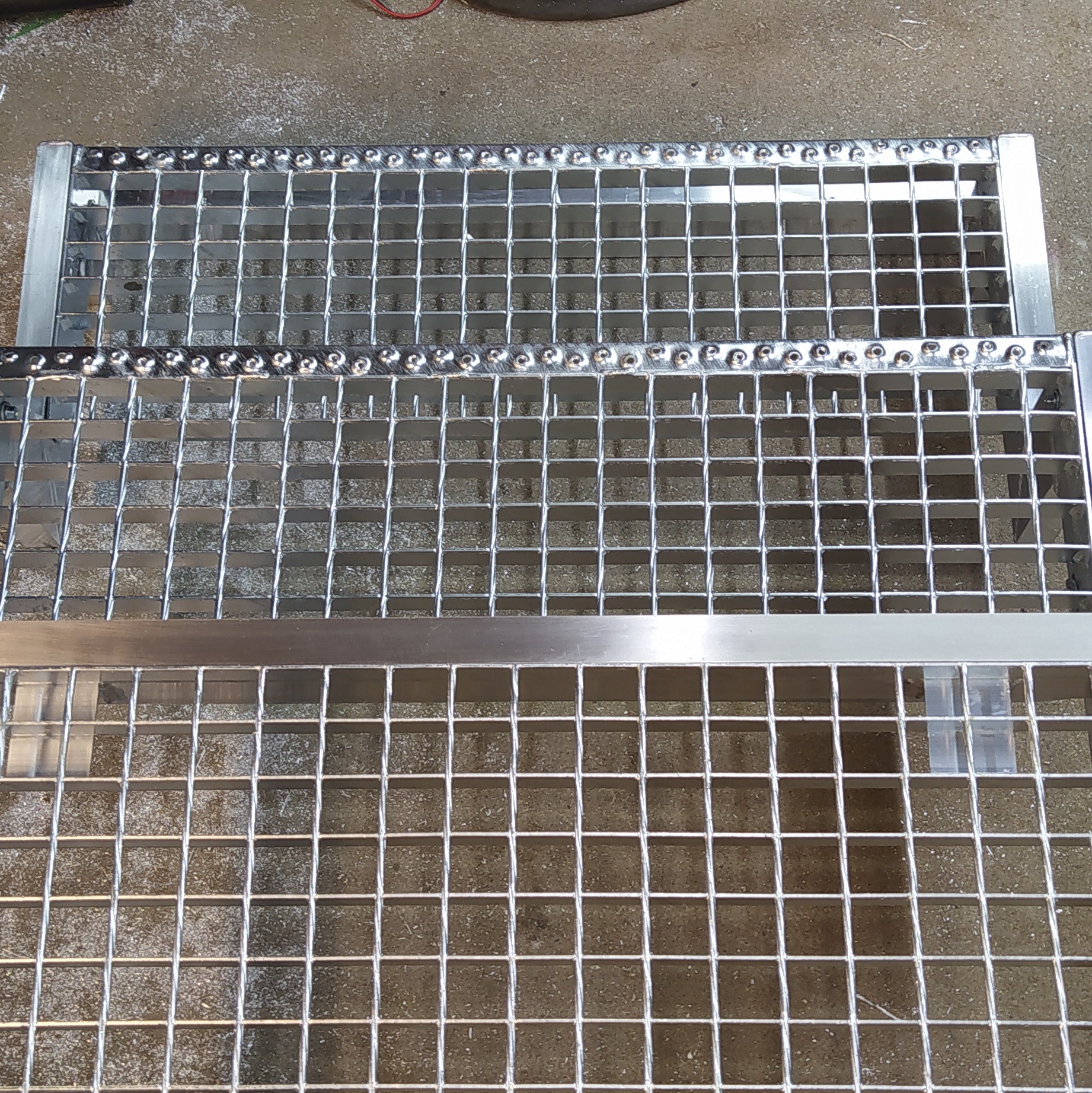 Metalowe kraty pomostowe z widocznymi spawami i nitami, ułożone jedna na drugiej, ukazujące precyzję wykonania i solidność konstrukcji.