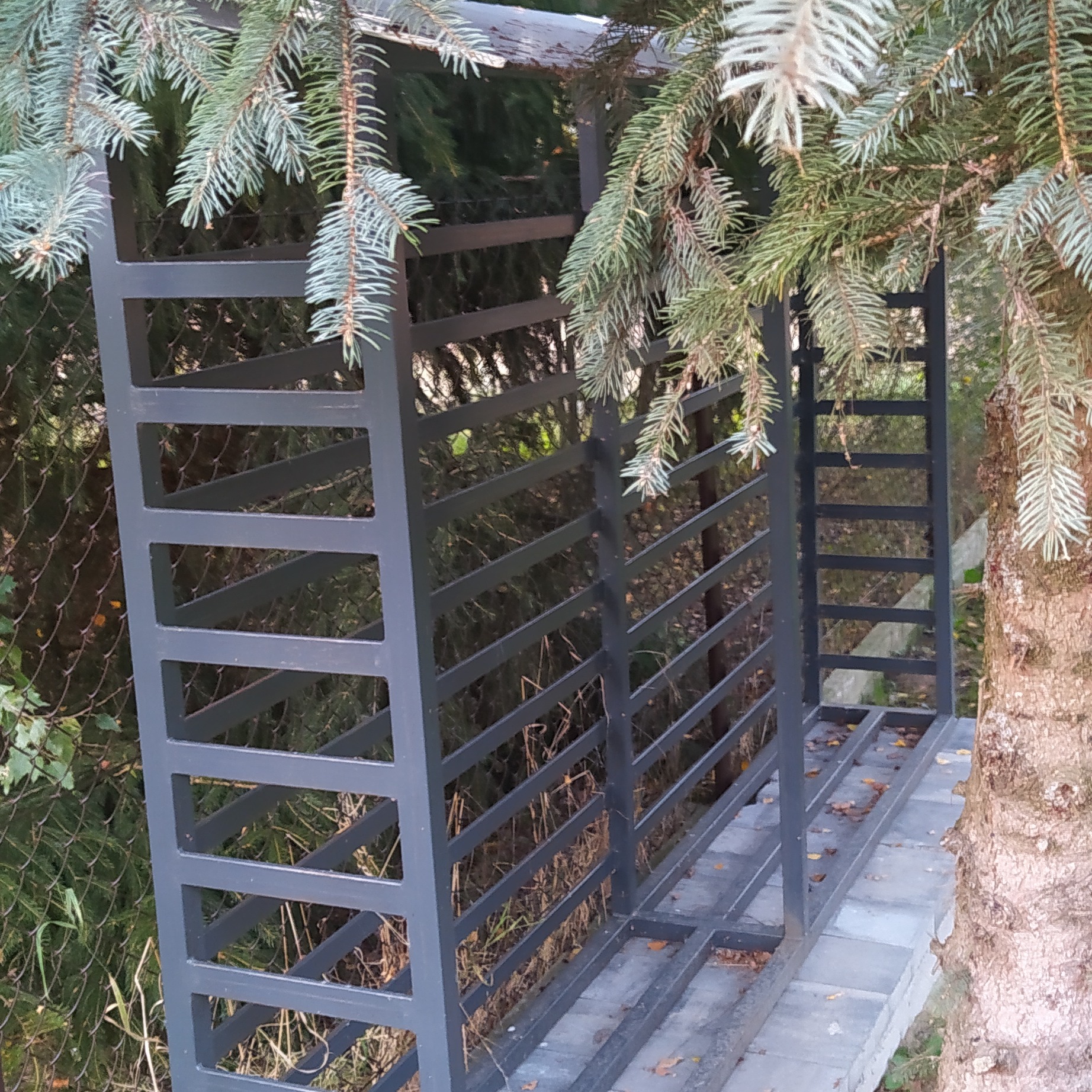 Metalowa konstrukcja, prawdopodobnie regał na drewno, pomalowana na szaro, umieszczona w ogrodzie, obok drzewa iglastego. Widoczne elementy spawane.