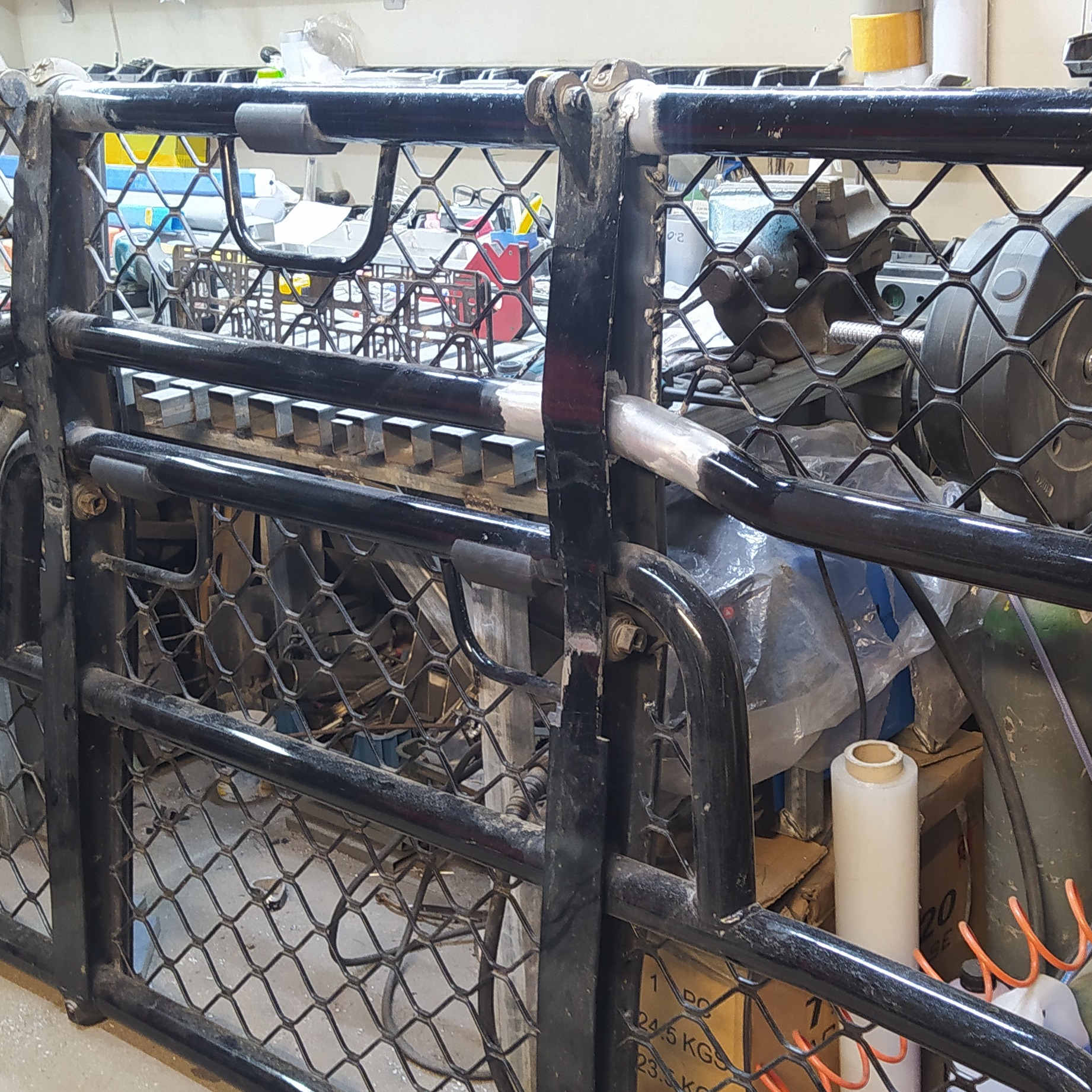 Metalowa konstrukcja z siatką, czarne rury i spawane elementy. Widoczne ślady obróbki, surowy wygląd. W tle warsztat z narzędziami i materiałami.