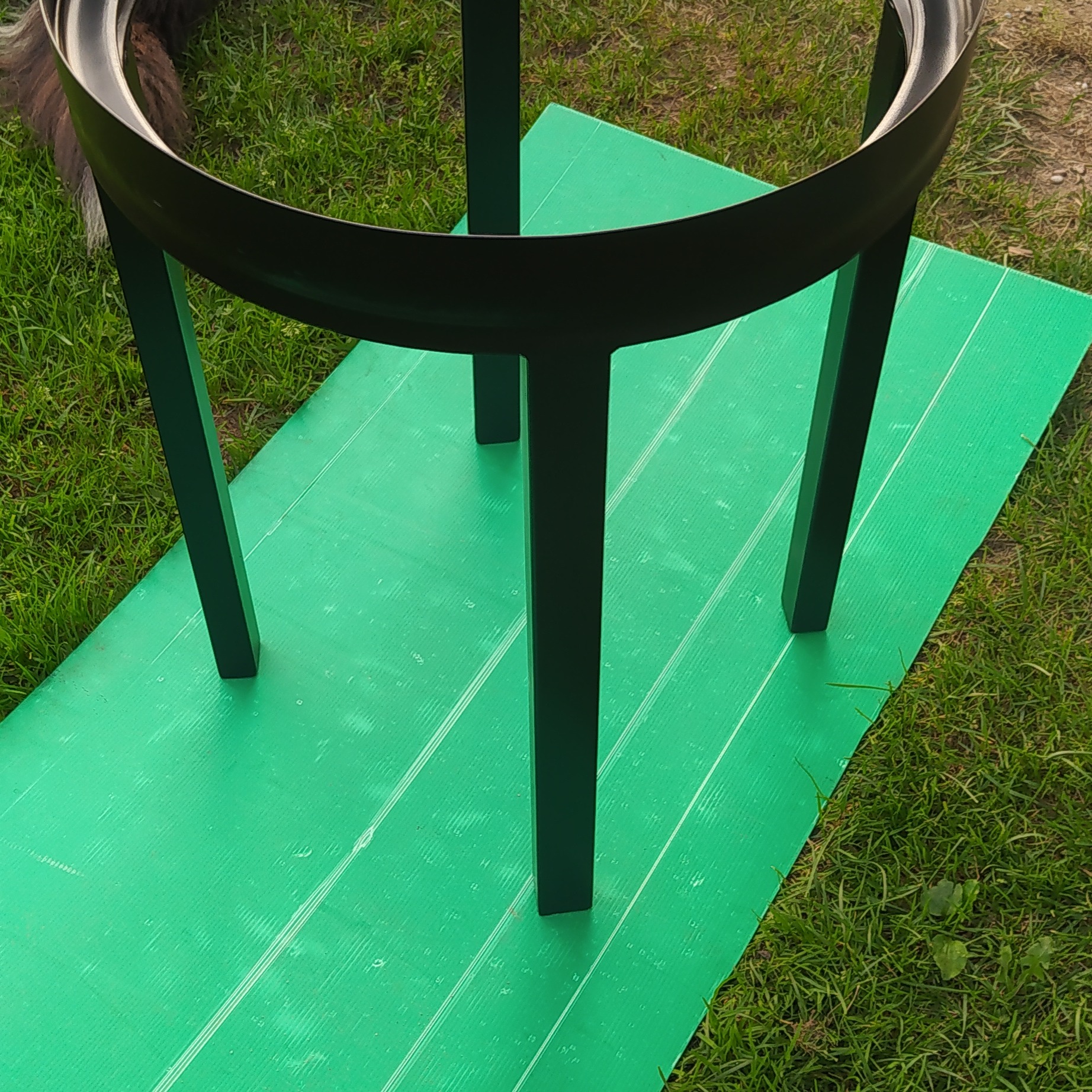 Czarna, metalowa konstrukcja z okrągłym elementem na trzech nogach, ustawiona na zielonej, karbowanej macie na trawie.