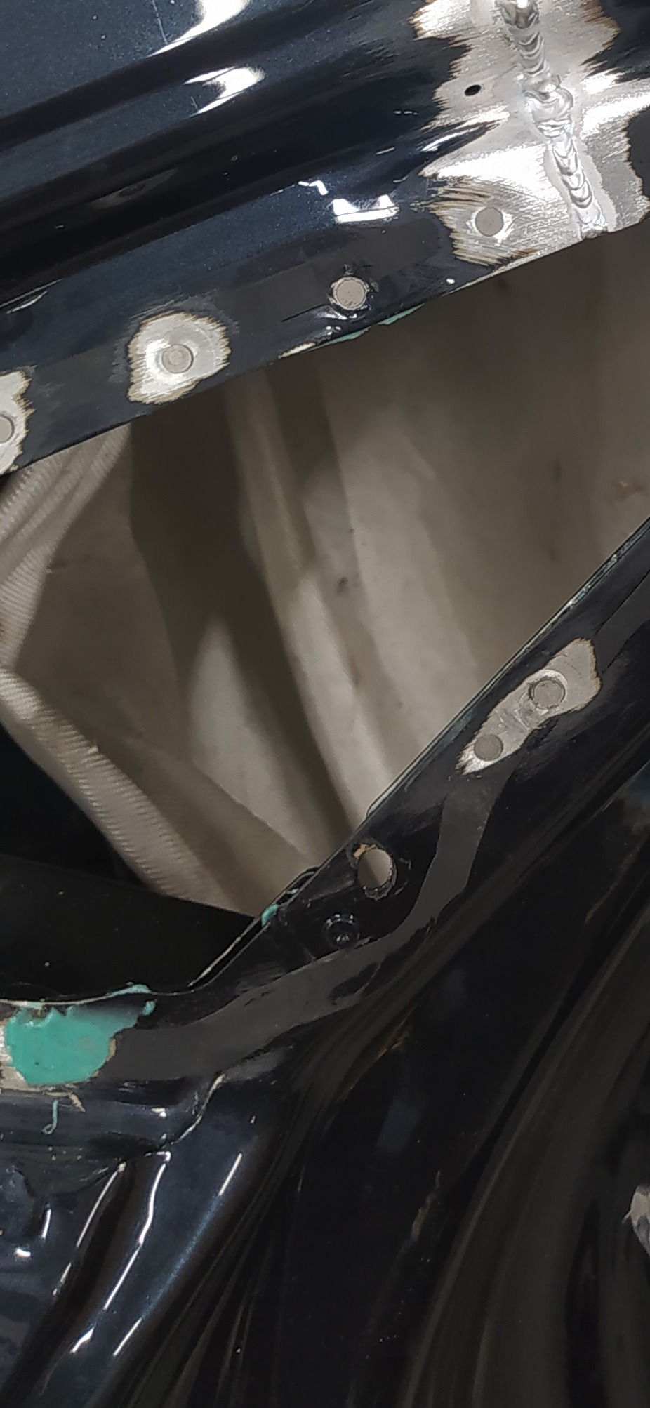 Detal spawu punktowego na czarnej karoserii samochodu, widoczne ślady szlifowania i otwory montażowe. Tkanina w tle. Precyzyjne łączenie blach.