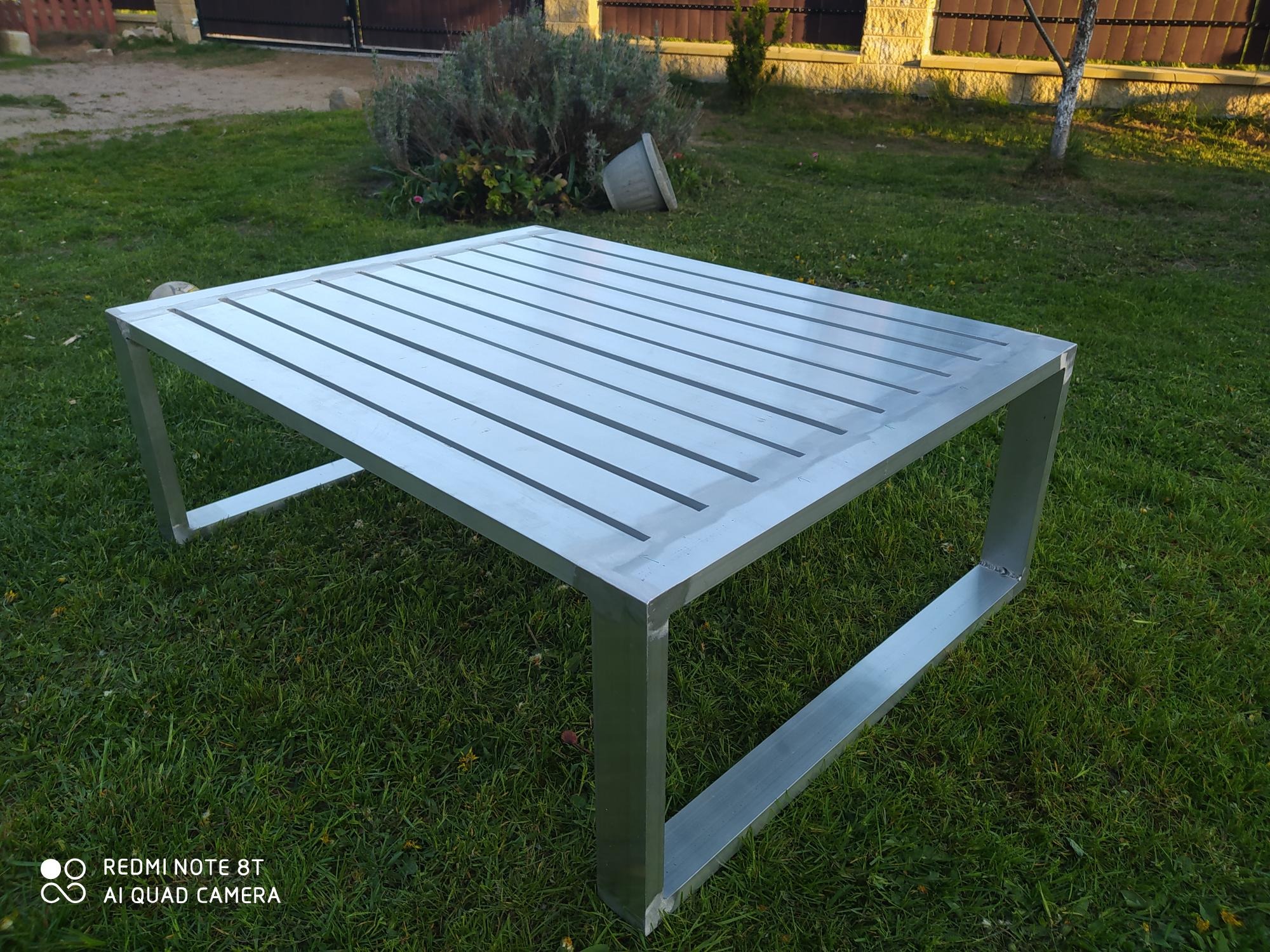 Stół z profili aluminiowych, spawany, z ażurowym blatem, ustawiony na trawie. Widoczne spawy w narożnikach. Tło: trawnik, krzewy i fragment ogrodzenia.