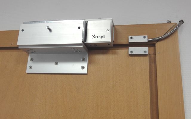 Zamontowany elektrozaczep marki Yotogi na drewnianych drzwiach, widoczne okablowanie w metalowej osłonie oraz elementy mocujące.