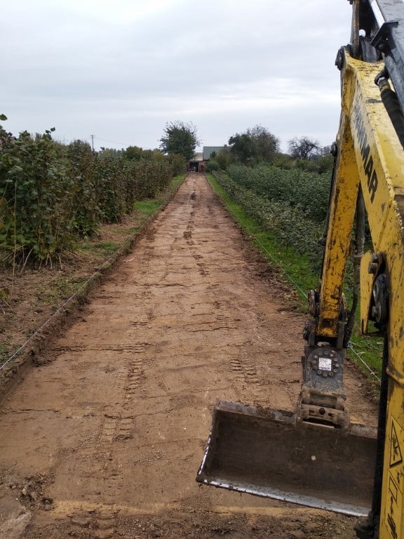 Żółta koparka Yanmar częściowo widoczna z prawej strony kadru, wyrównuje teren przyszłej drogi gruntowej, widoczne ślady opon, po bokach szpalery krzewów, w tle budynek gospodarczy.