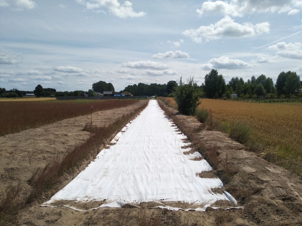 Biała geowłóknina rozłożona w wykopie wzdłuż pola, zabezpieczająca przed erozją, z widocznymi śladami po pracy sprzętu i wystającymi pomarańczowymi tyczkami.