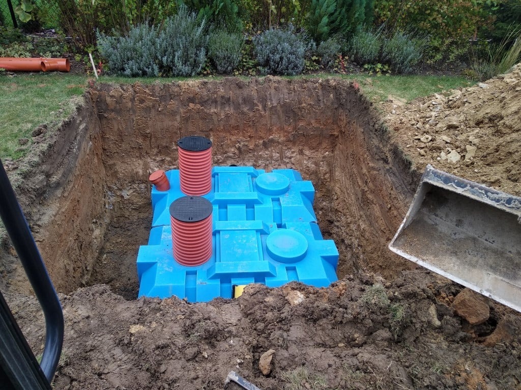 Niebieski zbiornik na deszczówkę z czerwonymi kominami inspekcyjnymi w wykopie o równych ścianach, widoczna łyżka koparki i rury na trawie.