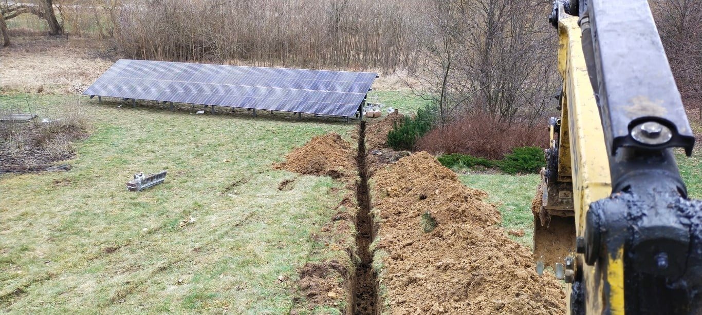 Długi wykop liniowy pod instalację, z widocznymi panelami słonecznymi w tle, wykonany koparką na trawiastym terenie.