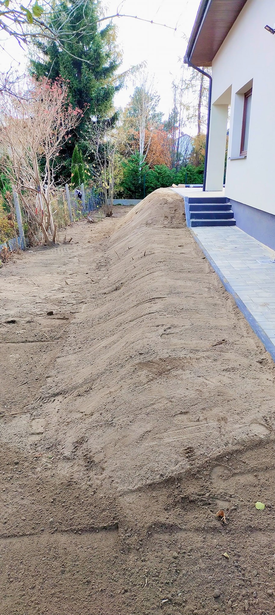 Przygotowany teren pod trawnik z wyrównaną ziemią, widoczny chodnik z kostki brukowej i schody prowadzące do domu.