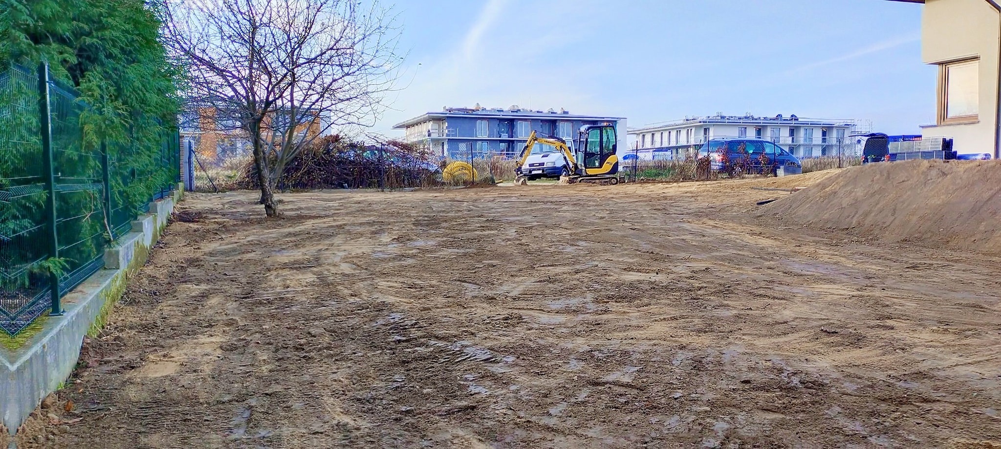 Żółta minikoparka stoi na placu budowy, w tle budynki mieszkalne i sterta gałęzi, teren przygotowany pod dalsze prace.