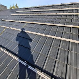 EKODOM24 SPÓŁKA Z OGRANICZONĄ ODPOWIEDZIALNOŚCIĄ - Montaż szyn pod panele fotowoltaiczne na grafitowym dachu z blachodachówki, cień osoby w lewym dolnym rogu. Widok z góry, słoneczny dzień.