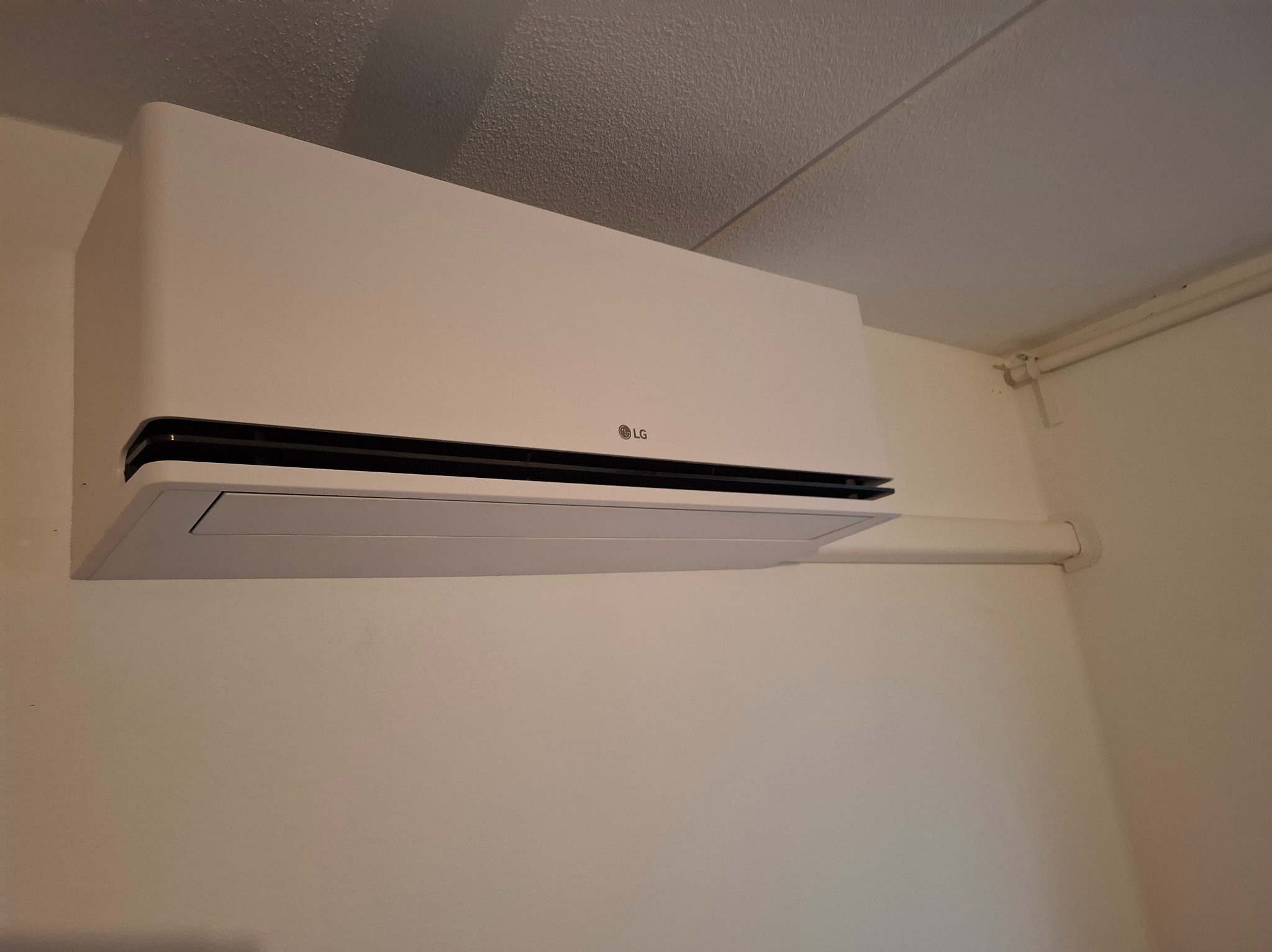 Biały klimatyzator LG zamontowany na ścianie w pomieszczeniu. Widoczne rury instalacyjne. Minimalistyczny design urządzenia.